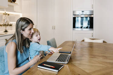 Boy pointing at laptop while sitting on mother's lap working in kitchen - XLGF01945