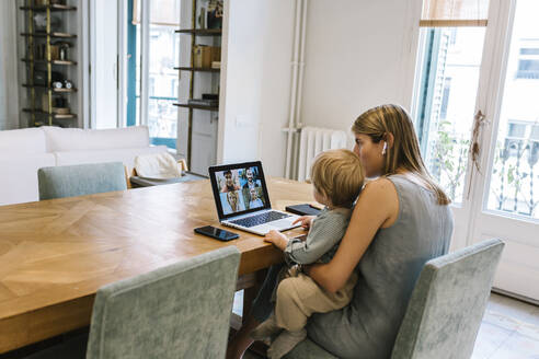 Boy sitting with mother during video conference through laptop at home - XLGF01946