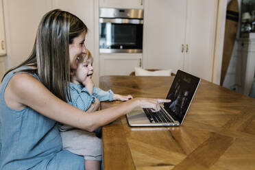 Smiling woman pointing at laptop while son sitting on lap during video call with family in kitchen - XLGF01950