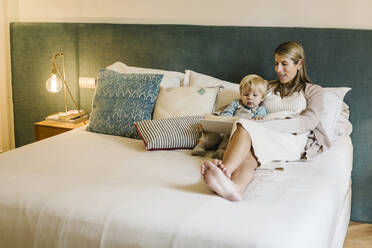 Pregnant woman talking with son while showing photo album on bed at home - XLGF01958