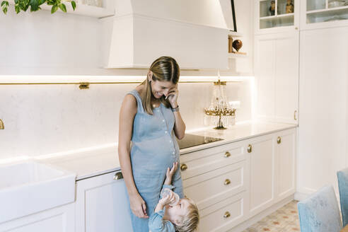 Smiling woman talking on mobile phone while looking at son drinking milk in kitchen - XLGF01968