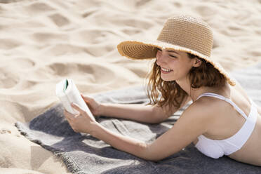 Lächelnde Frau mit Hut, die wegschaut, während sie in den Ferien auf einem Handtuch am Strand liegt - SBOF03854