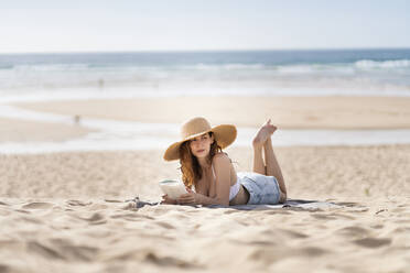 Junge Frau mit Buch, die wegschaut, während sie am Strand im Urlaub auf dem Sand liegt - SBOF03855
