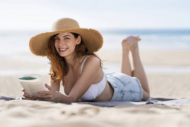 Lächelnde schöne Frau mit Buch liegt auf Handtuch am Strand während sonnigen Tag - SBOF03856