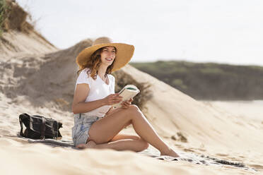 Lächelnde junge Frau mit Hut sitzt mit Buch an einem sonnigen Tag am Strand während der Ferien - SBOF03858