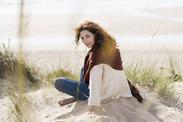 Lächelnde Frau schaut über die Schulter, während sie mit gekreuzten Beinen am Strand sitzt - SBOF03861