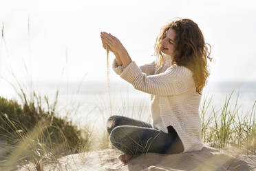 Smiling woman playing with sand amidst dunes on beach during vacations - SBOF03863