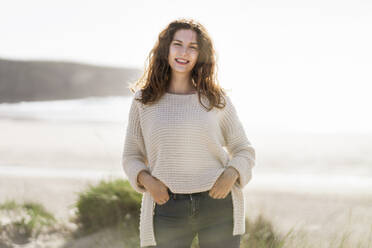 Smiling woman with hands in pockets standing at beach during sunny day - SBOF03865