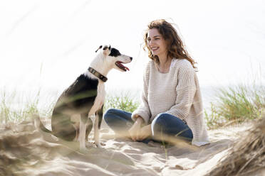 Lächelnde Frau sieht ihren Hund an, während sie im Urlaub im Sand sitzt - SBOF03868