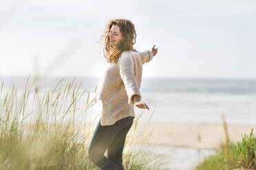 Carefree woman with arms outstretched standing amidst dunes at beach - SBOF03869