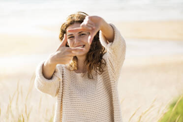 Happy woman looking through finger frame at beach during sunny day - SBOF03870