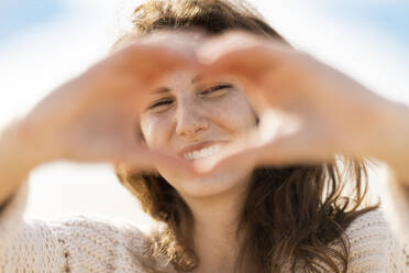 Glückliche junge Frau, die durch ein von Hand geformtes Herz am Strand während eines sonnigen Tages schaut - SBOF03872