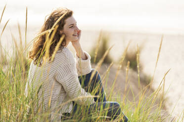 Young woman with hand on chin contemplating while sitting in dunes at beach during vacations - SBOF03874