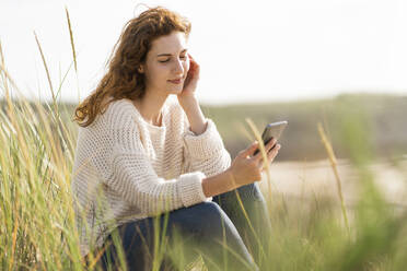 Young woman using mobile phone in dunes on beach during sunny day - SBOF03875