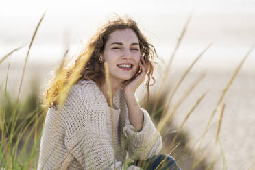 Smiling young woman with hand on chin sitting in dunes at beach - SBOF03878