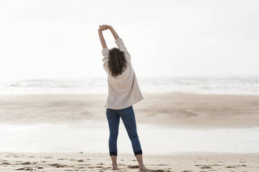 Junge Frau mit erhobenen Armen und Blick auf die Aussicht, während sie am Strand steht - SBOF03887