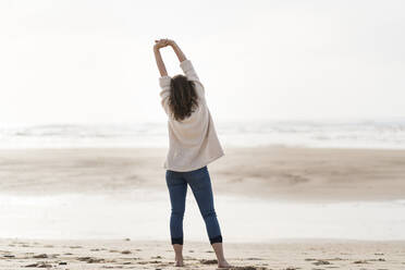 Young woman with arms raised hands looking at view while standing at beach - SBOF03887