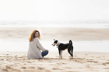 Frau schaut dem Hund über die Schulter, während sie im Urlaub am Strand sitzt - SBOF03889