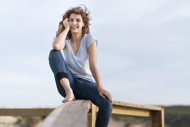 Smiling woman with head in hand sitting on railing in front of sky - SBOF03894