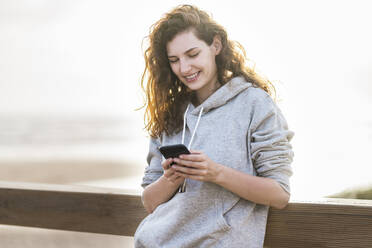 Smiling woman using smart phone while leaning on railing during sunny day - SBOF03896