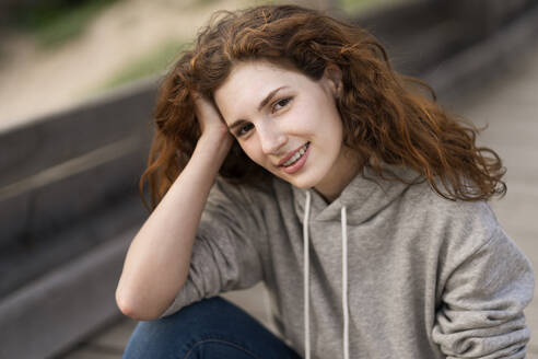 Lächelnde schöne Frau mit Hand im Haar auf einer Brücke sitzend - SBOF03901