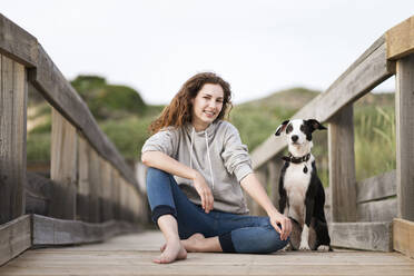 Smiling young woman sitting by Jack Russell Terrier dog on wooden bridge - SBOF03902