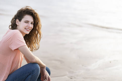 Lächelnde junge Frau, die am Strand im Sand hockt - SBOF03904