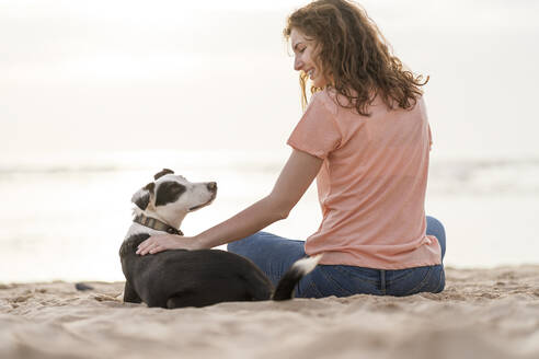 Lächelnde junge Frau, die einen Jack Russell Terrier streichelt, während sie am Strand sitzt - SBOF03909