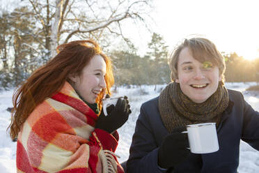 Smiling woman drinking coffee wrapped in blanket looking at boyfriend - FVDF00213