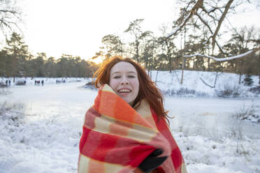 Fröhliche rothaarige Frau in Decke eingewickelt im Winter bei Sonnenuntergang - FVDF00221