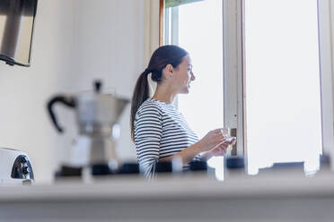 Junge Frau schaut durch ein Fenster und hält eine Kaffeetasse in der Küche - EIF01065