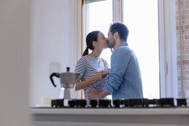 Couple kissing by window in kitchen at home - EIF01066