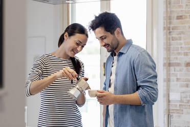 Lächelnde Frau gießt Kaffee ein, während der Mann eine Tasse in der Küche zu Hause hält - EIF01068