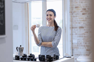 Glückliche junge Frau hält eine Kaffeetasse in der Hand, während sie in der Küche zu Hause steht - EIF01071