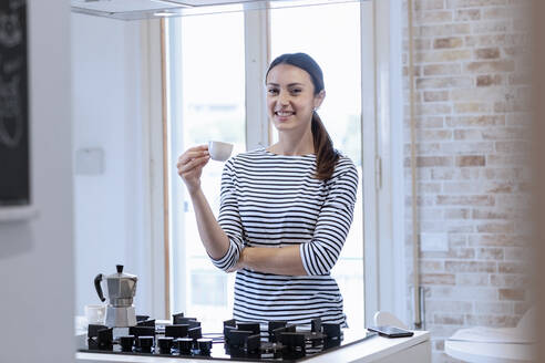 Happy young woman holding coffee cup while standing in kitchen at home - EIF01071