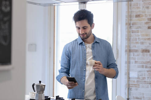Junger Mann benutzt ein Mobiltelefon und hält eine Kaffeetasse in der Küche - EIF01072