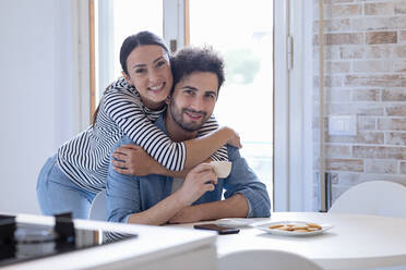 Smiling girlfriend embracing boyfriend with coffee cup in kitchen at home - EIF01075