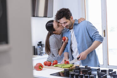 Young woman kissing boyfriend on cheek while cutting bell pepper in domestic kitchen - EIF01084