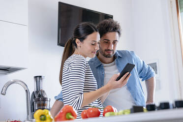 Lächelndes Paar, das in der heimischen Küche gemeinsam ein Mobiltelefon benutzt - EIF01086