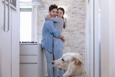 Lächelndes, sich umarmendes Paar mit Blick auf den Hund in der heimischen Küche - EIF01091