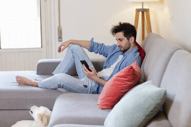 Mittelgroßer erwachsener Mann, der ein Mobiltelefon benutzt, während er zu Hause auf dem Sofa sitzt - EIF01097