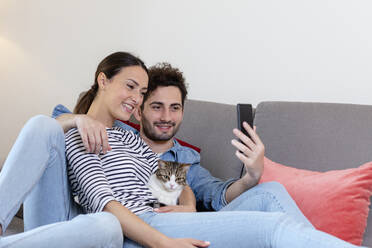Smiling couple with cat taking selfie through smart phone at home - EIF01101
