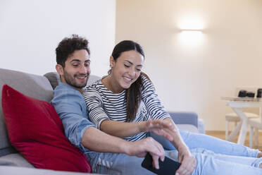 Happy couple laughing and playing with mobile phone on sofa at home - EIF01106