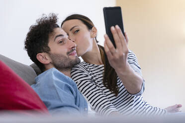 Freundin nimmt Selfie durch Smartphone beim Küssen auf Freund Wange zu Hause - EIF01109