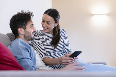 Smiling couple with smart phone sitting on sofa at home - EIF01110