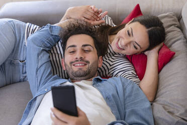 Happy couple using mobile phone while lying on sofa at home - EIF01119
