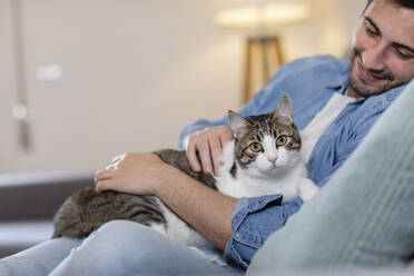 Smiling man stroking cat while sitting on sofa at home - EIF01140