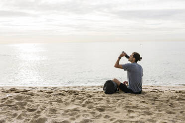 Junger Sportler trinkt Wasser, während er am Strand sitzt - XLGF01972