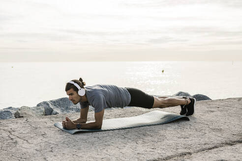 Männlicher Sportler mit Kopfhörern, der auf einer Trainingsmatte die Plank-Position übt - XLGF01976