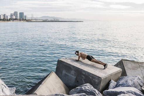 Young male athlete doing push-ups on block shape rock by sea - XLGF01982